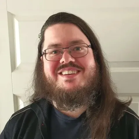 A picture of me, a long-haired person with a full beard, wearing glasses, stading in front of a white door.