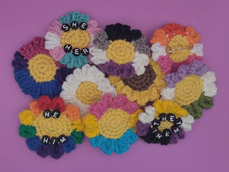 Crochet flower pins stacked on top of each other, shaped and colored like a daisy, a sunflower, or in various LGBTQIA+ pride flag colors. Some have letter beads spelling out pronouns. One pin is turned around, showing the brooch pin on its back.