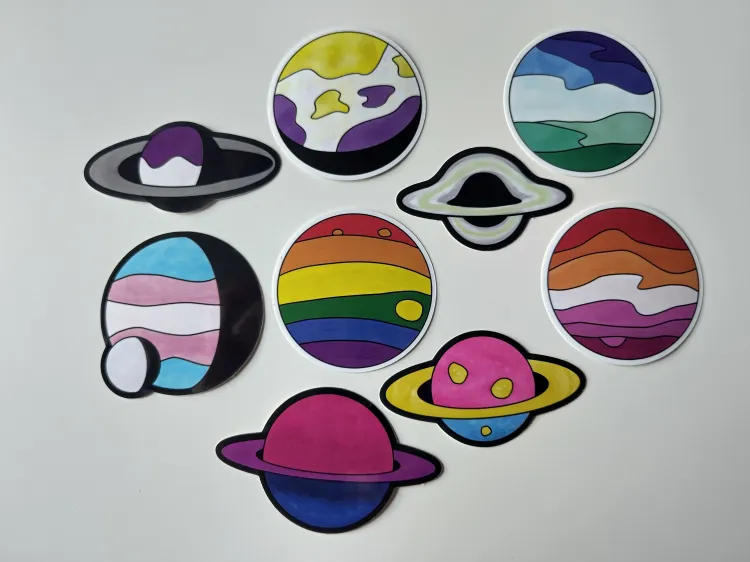 Nine colorful stickers on a white table. Eight depict planets, one an accreting black hole. Three planets have rings and one has a small moon. The colors match LGBTQIA+ pride flags: asexual, non-binary, MLM, trans, rainbow, agender, lesbian, bi, pan.