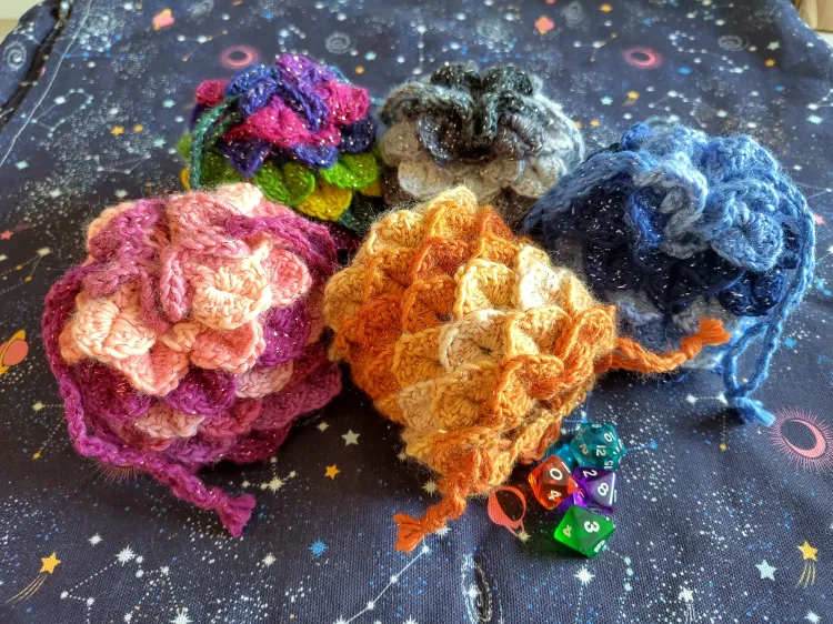 Five dice bags resembling scaly dragon eggs, in various colors. Four are closed, the fifth is open and lies on its side with dice spilling out.