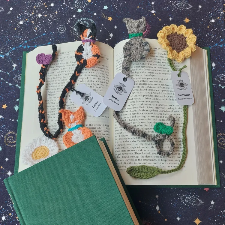Bookmarks depicting cats and flowers, placed in or on a book. Inside a closed green book, there are a daisy and an orange and white cat. On top of an open book are a black, white, and orange cat, a grey striped cat, and a sunflower.