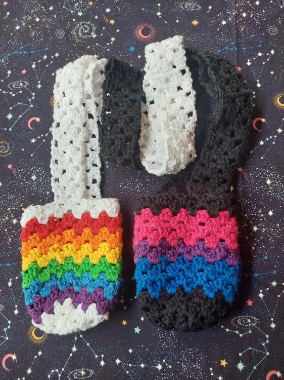 Two crocheted bottle holders laying flat. The were first worked as a cylinder and then as a flat strap. The left one is mostly white with rainbow colors in the cylinder. The right one is mostly black with stripes in blue, purple, and pink on the cylinder.