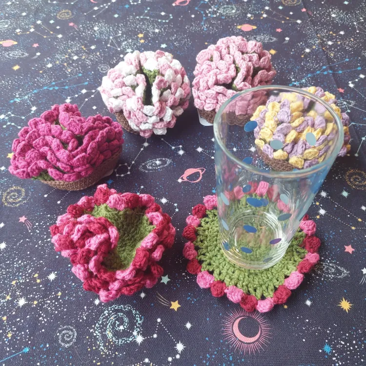 Five sets of crochet coasters that are green in the center and have colorful (e.g. purple, pink, yellow) petals on the outside. Groups of four coasters are placed in a brown crochet pots to look like a flower. One coaster is laid flat with a glass on top.