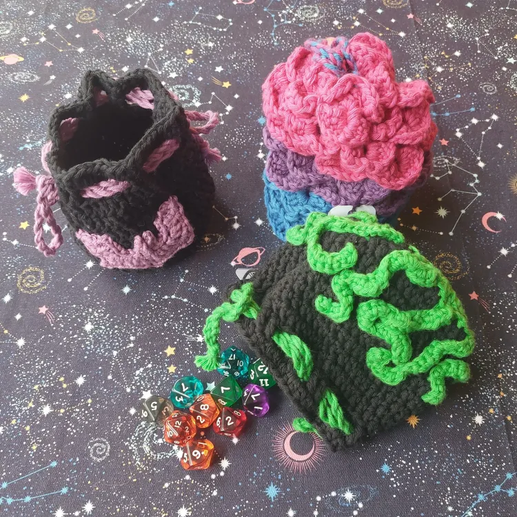 Three crochet dice bags. One is black with a pink bat appliqué, another has scales like a dragon in pink, purple, and blue. The last one is black with green tentacles and is laying on its side with dice spilling out.