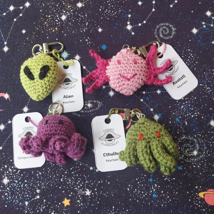 Four crochet keychain. From top left to bottom right: an light green alien head with black almond-shaped eyes, a pink axolotl, a purple octopus, the head of a green creature with tentacles in its face and red eyes.