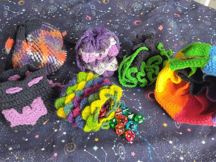 Six crochet dice bags in various styles and colors, includes dragon scales, an appliqué bat or tentacles, a color changing pattern, and one bag with multiple segments in the colors of the reainbow and internal pockets.