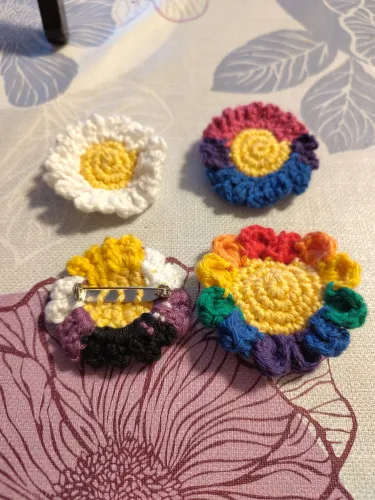 Four flower pins, all with a yellow center: a daisy and 18 white petals, one with 20 petals in pink, purple, and blue (bisexual pride), one with 16 petals in yellow, white, purple and black (non-binary pride), and one with 12 petals in 6 rainbow colors.