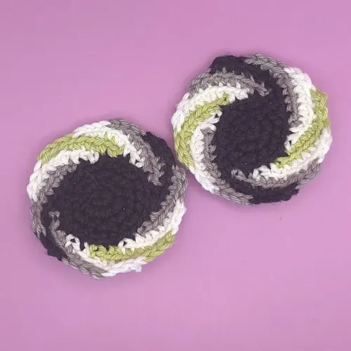 Two crochet pins with a black center and spiral arms in black, grey, white, and light green, the colors of the agender pride flag. The left pins has a larger black center than the right one.