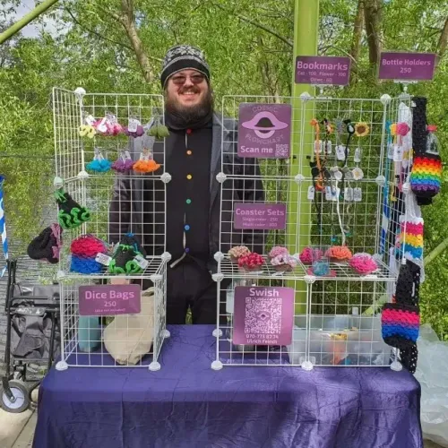 Me, a bearded person with darkened glasses, behind a table displaying my crochet items, including keychains, dice bags, coaster set, bookmarks, and bottle holders. Additionally, there are purple signs with the prices, my logo, and a QR code for payment.