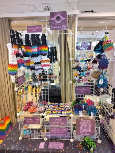 My setup for Queercon, a table displaying my crochet items, including keychains, pins, dice bags, coaster set, bookmarks, and bottle holders. Additionally, there are purple signs with the prices, my logo, and a QR code for payment.