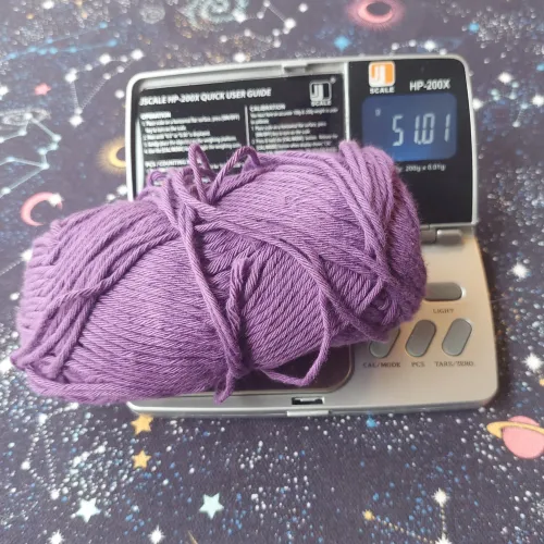 A ball of purple yarn laying on a small digital scale reading 51.01 grams, placed on a cloth depicting a starry sky with stars, planets, crescent moons, galaxies and constellations.