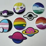Nine colorful stickers on a white table. Eight depict planets, one an accreting black hole. Three planets have rings and one has a small moon. The colors match LGBTQIA+ pride flags: asexual, non-binary, MLM, trans, rainbow, agender, lesbian, bi, pan.