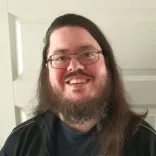 A picture of me, a long-haired person with a full beard, wearing glasses, stading in front of a white door.