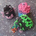 Three crochet dice bags. One is black with a pink bat appliqué, another has scales like a dragon in pink, purple, and blue. The last one is black with green tentacles and is laying on its side with dice spilling out.