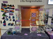 My setup for Retromania, a table displaying my crochet items, including keychains, pins, dice bags, coaster set, bookmarks, and bottle holders. Additionally, there are purple signs with the prices, my logo, and a QR code for payment.